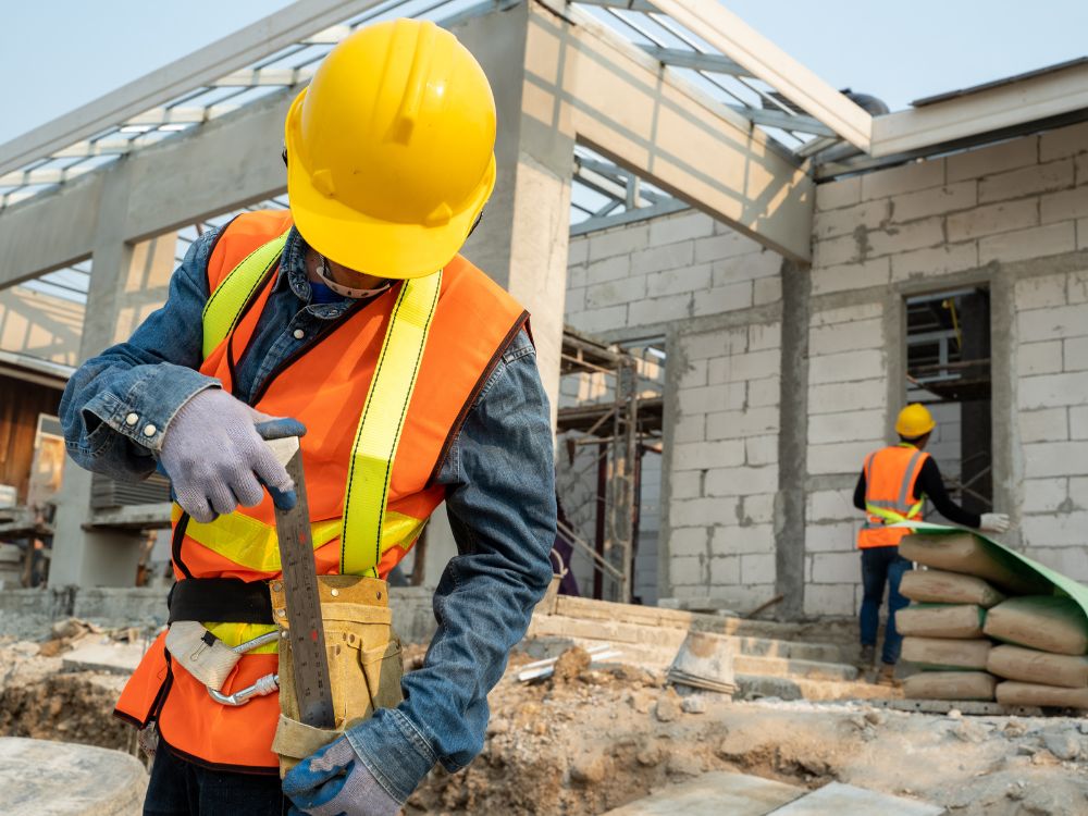 Trabalhador da construção civil usando EPI e ferramentas mede material em obra residencial, representando etapa de execução planejada da reforma.