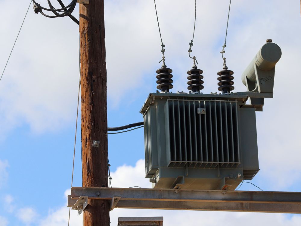 Um transformador elétrico cinza preso a um poste de madeira com fios e isoladores. O céu é azul com nuvens brancas e o transformador tem a grade de ventilação visível, mostrando sua estrutura robusta.