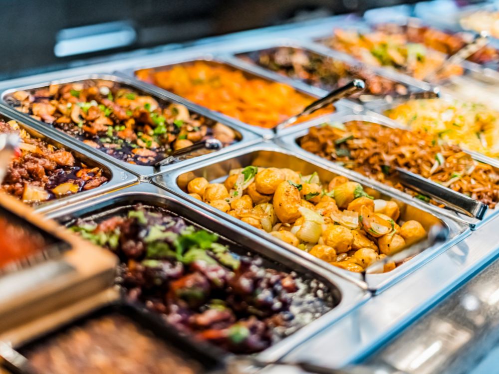 Buffet variado de comidas para o dia a dia com opções como batatas assadas, massas, carnes e legumes em recipientes de inox.