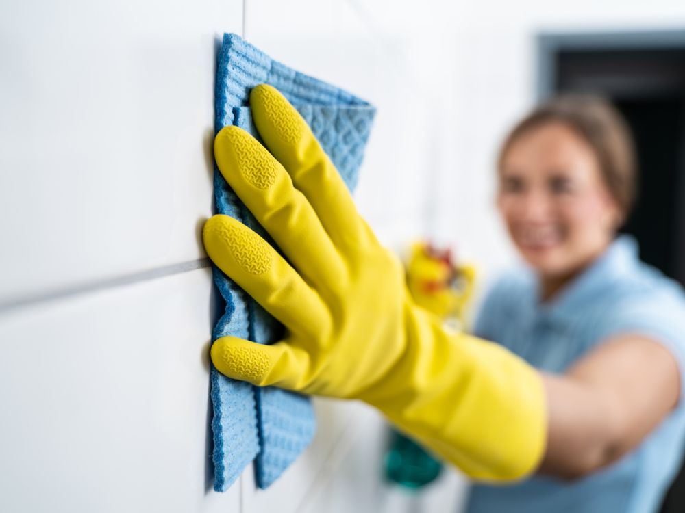 Pessoa utilizando luvas amarelas limpando rejunte de azulejos com pano, mostrando a higienização de revestimento no banheiro.