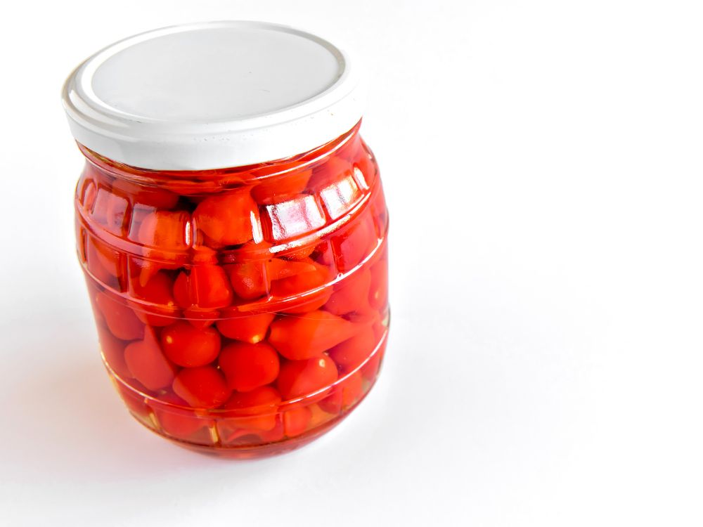 Pote de vidro com conserva de pimenta biquinho em líquido avermelhado sobre fundo branco, ilustrando como fazer conserva de pimenta biquinho de forma caseira.