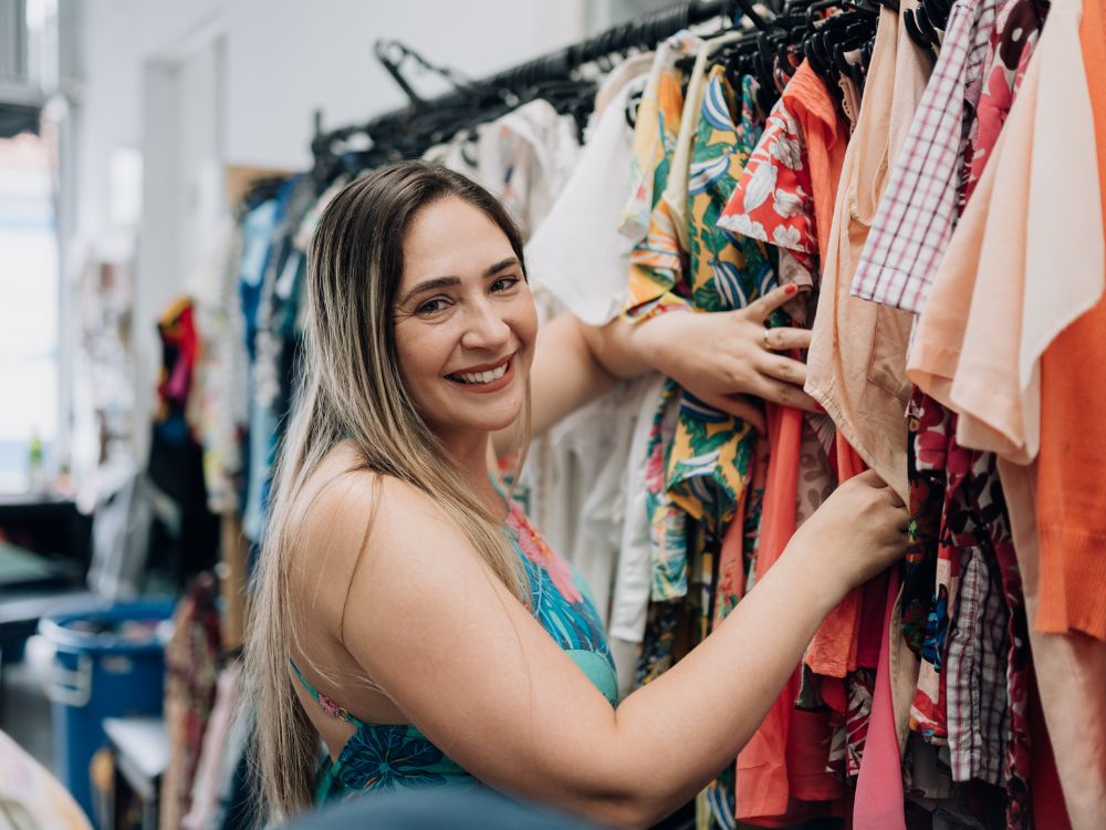 Mulher sorrindo organizando peças em uma arara de roupas coloridas dentro de loja de moda, ilustrando o custo para abrir loja de roupas e estruturar um espaço comercial atrativo.